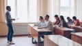 Male professor in casual clothes is talking to group of students sitting at tables in classroom and making notes. Large Royalty Free Stock Photo