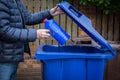 A male recycler placing a plastic bottle into a blue roadside recycling bin