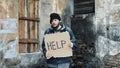 Homeless holds piece of cardboard with inscription Help. Refugee at abandoned building looking for job. Male tramp in dirty