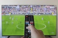 a male right hand holding a black TV remote control with a blurred football game in a smart tv