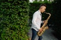 Male saxophonist plays on the alley in summer park