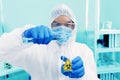 Male Scientist In PPE Suit Working In Lab While Pouring Solution From Cylinder To Erlenmeyer Flask With BioHazard Sign.