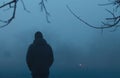 Male silhouette on background of the evening park in dense fog. Thick mist in dark scary place. Dark noir person and