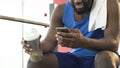 Male sitting in gym, holding protein cocktail and mobile phone, social newsfeed