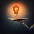 Male student hand holding an open book with a shining light bulb over a dark blackboard background. Symbol of knowledge, Royalty Free Stock Photo