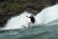 A male surfer in a black wetsuit rides a wave in front of cliffs at Piha Beach, Piha, Auckland, New Zealand