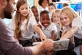 Male Teacher Reading Story To Group Of Elementary Pupils Wearing Uniform In School Classroom