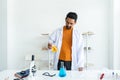 A male teacher in white lab coat with safety glasses putting blue chemical flask on table with many testing tools