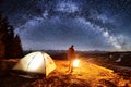 Male tourist have a rest in his camp near the forest at night. Man near campfire and tent under night sky full of stars Royalty Free Stock Photo