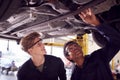 Male Tutor With Student Looking Underneath Car On Hydraulic Ramp On Auto Mechanic Course