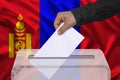 Male voter drops a ballot in a transparent ballot box on the background of the national flag of Mongolia, concept of state