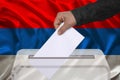 Male voter drops a ballot in a transparent ballot box on the background of the national flag of Serbia, concept of state elections