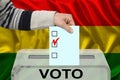 Male voter drops a ballot in a transparent ballot box against the background of the national flag of bolivia, concept of state
