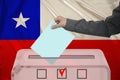 Male voter drops a ballot in a transparent ballot box on the background of the national flag of Chile, concept of state elections