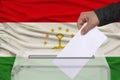 Male voter lowers the ballot in a transparent ballot box against the background of the national flag of Tajikistan, concept of