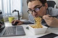 Male worker busy working with laptop, use chopsticks to hastily eat instant noodles during office lunch& x27;s break Royalty Free Stock Photo