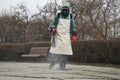 A male worker in a chemical protective suit sprays disinfecting liquid on a city street or in a park during quarantine and a