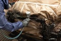 A male worker in jumpsuit and gloves paints with a spray gun a front frame part of the car body in black after being damaged at an