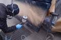 A male worker in jumpsuit and gloves paints with a spray gun a front frame part of the car body in black after being damaged at an