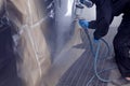 A male worker in jumpsuit and gloves paints with a spray gun a front frame part of the car body in black after being damaged at an
