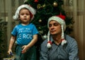 A little boy wearing Santa Claus hat and his uncle wearing Snow Maiden hat sitting on background of New Year tree.