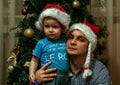 A little boy wearing Santa Claus hat and his uncle wearing Snow Maiden hat sitting on background of New Year tree.