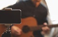 Man with acoustic guitar in a shirt and cap plays live staying home for the audience in front of the smartphone camera during