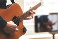 Man with acoustic guitar in a shirt and cap plays live staying home for the audience in front of the smartphone camera during