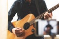 Man with acoustic guitar in a shirt and cap plays live staying home for the audience in front of the smartphone camera during