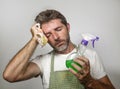 Man in apron holding kitchen cloth and detergent spray feeling overwhelmed and bored doing domestic housework of cleaning and