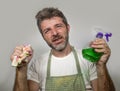 Man in apron holding kitchen cloth and detergent spray feeling overwhelmed and bored doing domestic housework of cleaning and