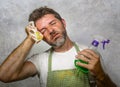 Man in apron holding kitchen cloth and detergent spray feeling overwhelmed and bored doing domestic housework of cleaning and