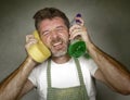 Man in apron holding sponge and detergent spray feeling overwhelmed and bored doing domestic housework of cleaning and washing