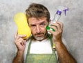 Man in apron holding sponge and detergent spray feeling overwhelmed and bored doing domestic housework of cleaning and washing in