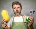 Man in apron holding sponge and detergent spray feeling overwhelmed and bored doing domestic housework of cleaning and washing in