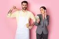 Man in apron holding wooden rolling pin near businesswoman with money talking on smartphone on pink