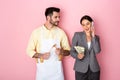 Man in apron holding wooden spoons and looking at businesswoman with money talking on smartphone on pink