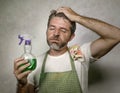 Man in apron with kitchen cloth and detergent spray feeling overwhelmed and bored doing domestic housework of cleaning and washing