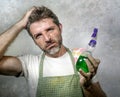 Man in apron with kitchen cloth and detergent spray feeling overwhelmed and bored doing domestic housework of cleaning and washing