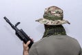 A man in an army panama hat and a military T-shirt holds a rifle with his back to the camera, a light background. Concept: soldier