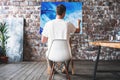 Man artist sitting on chair in front of canvas on easel and drawing. Painter working in workshop Royalty Free Stock Photo