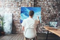 Man artist sitting on chair in front of canvas on easel and drawing. Painter working in workshop Royalty Free Stock Photo