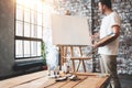 Man artist stands in front of a blank canvas on easel with palette in loft art studio. Male painter hold paintbrush in hand and mi