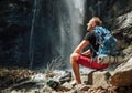 Muž s batohom oblečený v aktívnom trekingovom oblečení si vyzúva trekingové topánky, sedí pri vodopáde horskej rieky a užíva si