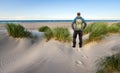 Man with Backpack hiking in beautiful windy coastal dune marram grass towards beach of North Sea in soft evening sunset