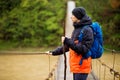 Man with backpack trekking in forest by hinged bridge over river. Cold weathe. Spring hiking. Wooden bridge across the river Royalty Free Stock Photo