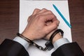 A man with bare hands in handcuffs sits at a table in front of a blank sheet of paper and a fountain pen. Concept: the detainee