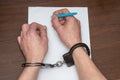 A man with bare hands in handcuffs sits at a table in front of a blank sheet of paper and a fountain pen. Royalty Free Stock Photo