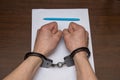 A man with bare hands in handcuffs sits at a table in front of a blank sheet of paper and a fountain pen. 2