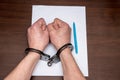 A man with bare hands in handcuffs sits at a table in front of a blank sheet of paper and a fountain pen. 4 Royalty Free Stock Photo